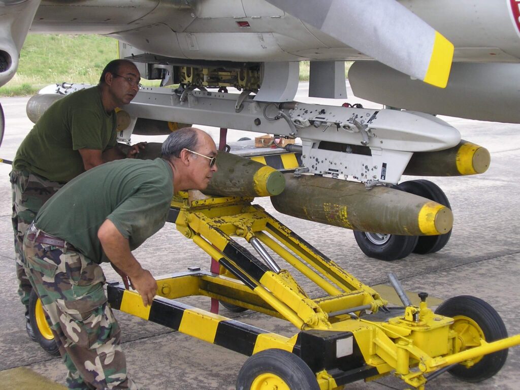 Weapons specialist (“armeros”) from Escuadrón 1 fit a 500 lb (250 kg) HE (High Explosive) bomb on a TER (Triple Ejector Rack) on a Pucará ventral hardened point. (Image Escuadrón 1)